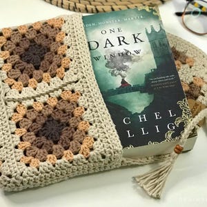 May include: A crocheted book sleeve with a brown and beige pattern. The sleeve is holding a book titled "One Dark Window" by Rachel Gillig.