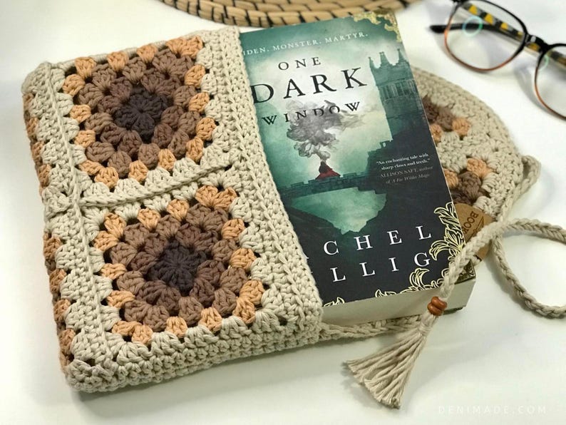 May include: A crocheted book sleeve with a brown and beige pattern. The sleeve is holding a book titled "One Dark Window" by Rachel Gillig.