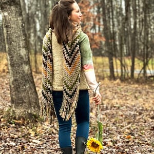Op de afbeelding: Een vrouw draagt een lange, gehaakte sjaal met een chevron-patroon in tinten groen, bruin en geel. De sjaal heeft franjes aan de uiteinden. Ze draagt een blauwe jeans en een cr&egrave;mekleurige trui. Ze loopt door een bosrijk gebied en houdt een zonnebloem vast.