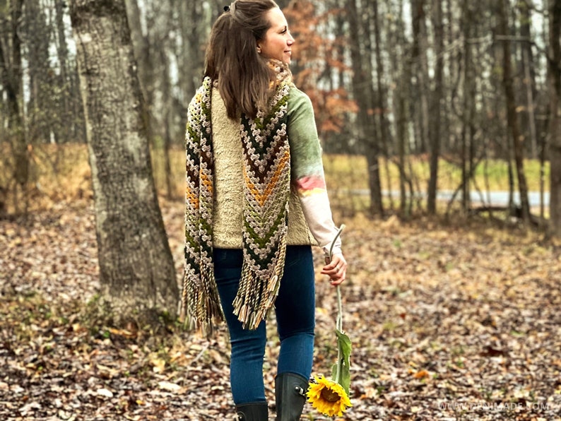 Op de afbeelding: Een vrouw draagt een lange, gehaakte sjaal met een chevron-patroon in tinten groen, bruin en geel. De sjaal heeft franjes aan de uiteinden. Ze draagt een blauwe jeans en een cr&egrave;mekleurige trui. Ze loopt door een bosrijk gebied en houdt een zonnebloem vast.