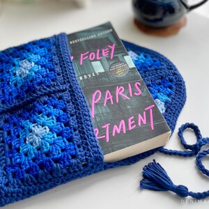 May include: A blue crocheted book sleeve with a book titled "Paris Apartment" by  Sarah Foley. The sleeve has a flap that folds over the book and a tassel.