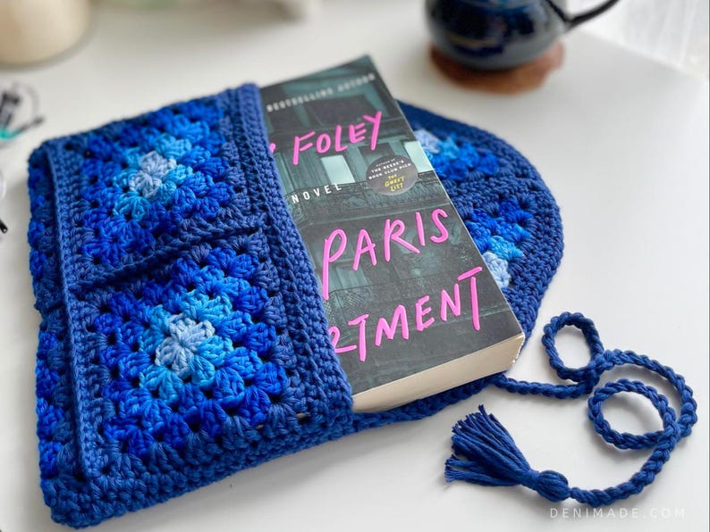 May include: A blue crocheted book sleeve with a book titled "Paris Apartment" by  Sarah Foley. The sleeve has a flap that folds over the book and a tassel.
