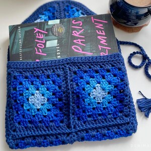 May include: A blue crocheted book sleeve with a tassel. The book sleeve has two large squares with a gradient of blue colors. The book sleeve is holding a book titled "Paris Apartment" by "S.J. Foley".