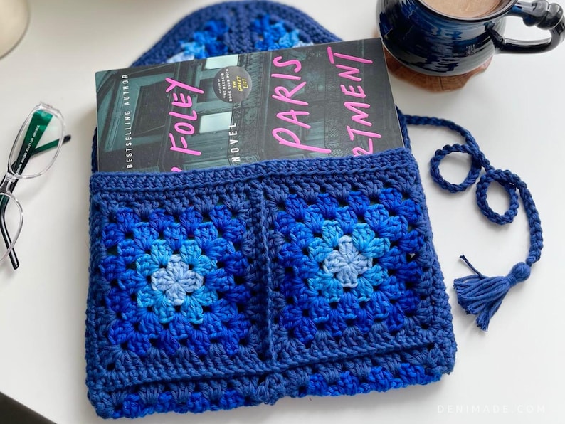May include: A blue crocheted book sleeve with a tassel. The book sleeve has two large squares with a gradient of blue colors. The book sleeve is holding a book titled "Paris Apartment" by "S.J. Foley".