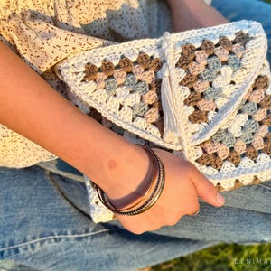 May include: A white crocheted clutch bag with a brown, pink, and blue granny square pattern. The bag has a white tassel and is held by a person wearing blue jeans.