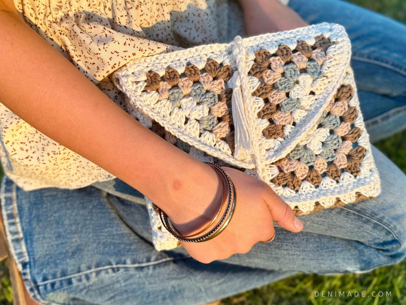 May include: A white crocheted clutch bag with a brown, pink, and blue granny square pattern. The bag has a white tassel and is held by a person wearing blue jeans.