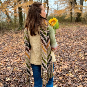 Op de afbeelding: Een vrouw draagt een bruin, groen en geel gehaakt sjaaltje met franjes, een cr&egrave;mekleurige trui en een blauwe spijkerbroek. Ze houdt een zonnebloem in haar hand en staat in een bos met gevallen bladeren op de grond.