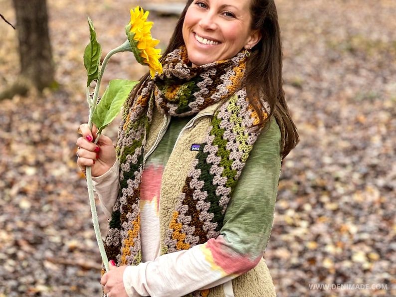 Op de afbeelding: Een vrouw draagt een groen shirt en een bruin, groen en oranje gebreide sjaal en houdt een gele zonnebloem vast.