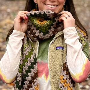 Op de afbeelding: Een bruine, groene en gele gehaakte sjaal met een granny square patroon. De sjaal wordt gedragen door een persoon met een groene fleece vest en een tie-dye shirt.