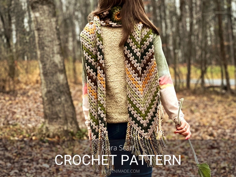 Op de afbeelding: Een vrouw draagt een lange, gehaakte sjaal met een chevron-patroon in tinten groen, bruin en beige. De sjaal heeft een franje aan de onderkant. De tekst "Klara Scarf CROCHET PATTERN WDENIMADE.COM" is zichtbaar onderaan de afbeelding.