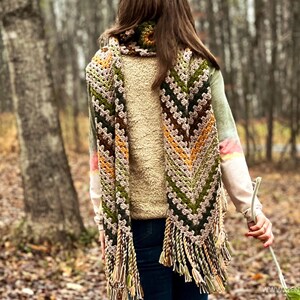 Op de afbeelding: Een gehaakte sjaal met een chevron-patroon in tinten groen, bruin, beige en oranje. De sjaal heeft franjes aan de uiteinden en wordt gedragen door een persoon in een bosrijke omgeving.
