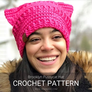 May include: A pink crocheted hat with cat ears. The hat has a brown leather patch on the side. The hat is being worn by a person with dark hair and a big smile. The text "Brooklyn Pussycat Hat CROCHET PATTERN WWW.DENIMADE.COM" is at the bottom of the image.