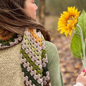 Op de afbeelding: Een vrouw draagt een gehaakte sjaal in tinten groen, bruin en geel. De sjaal is een granny square patroon. De vrouw houdt een gele zonnebloem vast.