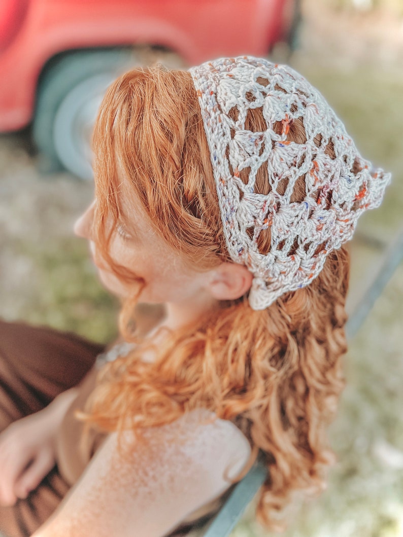 May include: A white crocheted headband with a lacy pattern and colorful flecks. The headband is worn by a person with long, curly red hair.