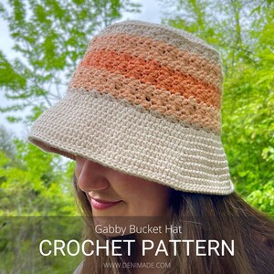Op de afbeelding: Een gehaakte bucket hat met een crème basis en oranje en perzikkleurige strepen. De hoed wordt gedragen door een persoon met lang bruin haar. De tekst "Gabby Bucket Hat CROCHET PATTERN www.denimade.com" wordt onder de hoed weergegeven.