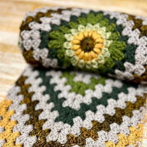 Op de afbeelding: Een gehaakt deken met een granny square patroon in tinten grijs, groen en geel.