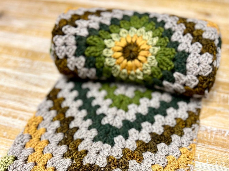 Op de afbeelding: Een gehaakt deken met een granny square patroon in tinten grijs, groen en geel.