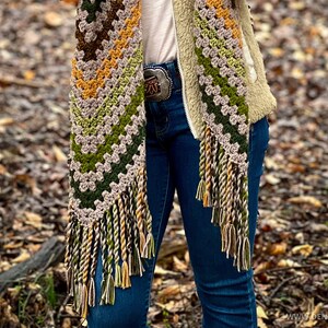 Op de afbeelding: Een gehaakte sjaal met franjes in tinten groen, bruin en geel. De sjaal wordt gedragen door iemand die een blauwe spijkerbroek en een bruin jack draagt.