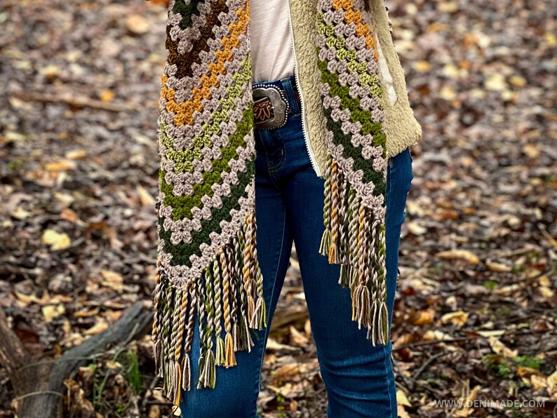 Op de afbeelding: Een gehaakte sjaal met franjes in tinten groen, bruin en geel. De sjaal wordt gedragen door iemand die een blauwe spijkerbroek en een bruin jack draagt.