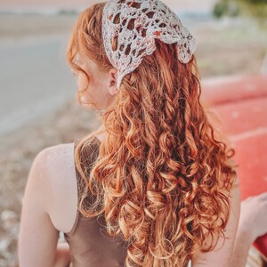 May include: A woman with long, curly, auburn hair is wearing a white crocheted headband with a floral pattern. The headband is tied at the back of her head. She is wearing a brown sleeveless dress.