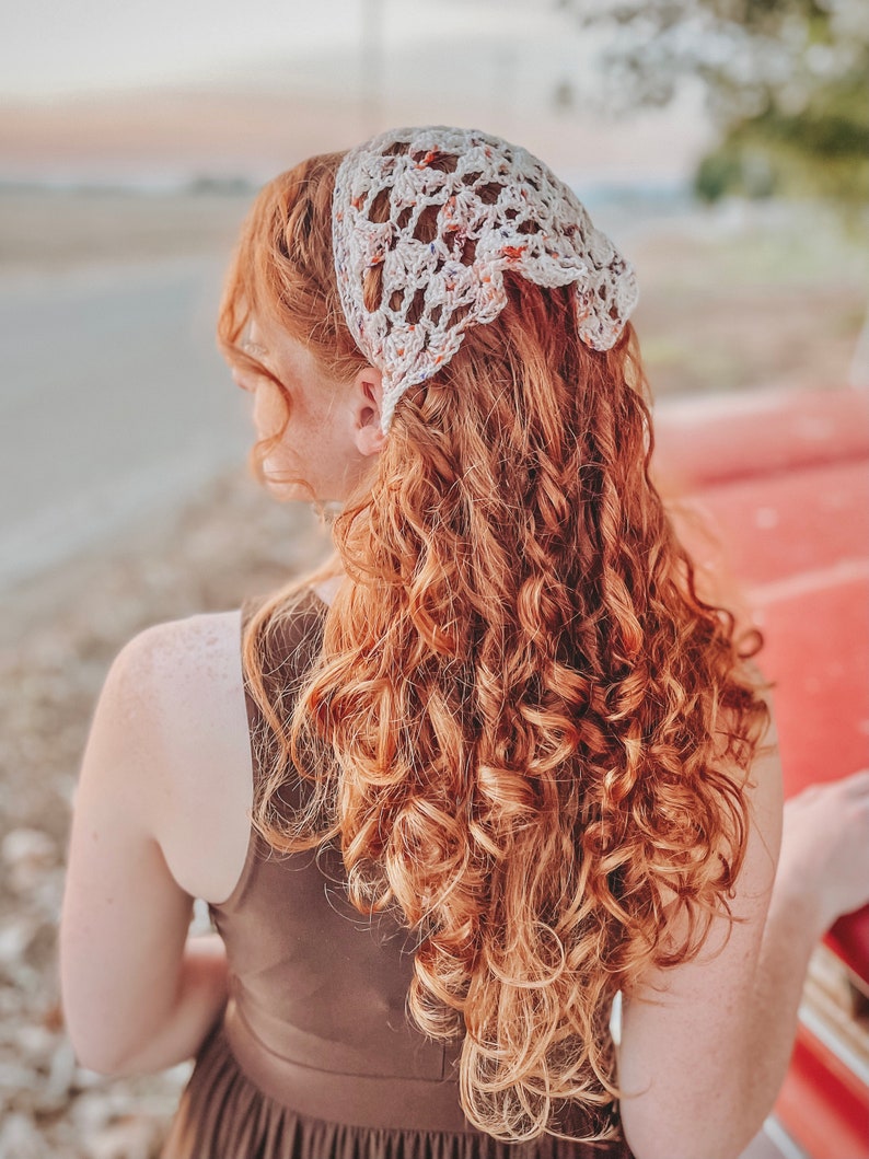May include: A woman with long, curly, auburn hair is wearing a white crocheted headband with a floral pattern. The headband is tied at the back of her head. She is wearing a brown sleeveless dress.