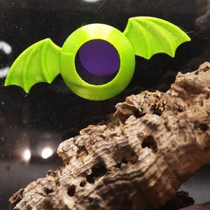 May include: A green plastic toy bat with a purple eye. The bat is sitting on a piece of brown bark.