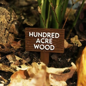 May include: A brown wooden sign with white text that reads "Hundred Acre Wood". The sign is propped up in the ground with a wooden stick.