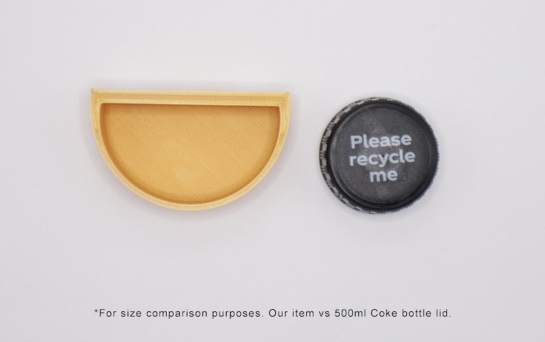 May include: A half-circle wooden tray next to a black plastic bottle cap with the text "Please recycle me". The tray is about the same size as a 500 ml Coke bottle lid.