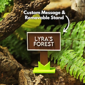 May include: A brown sign with the text "LYRA'S FOREST" in white lettering. The sign is on a brown post with a green base. The background includes green ferns and a tree branch. The text "Custom Message & Removable Stand" is also visible.