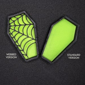 Op de afbeelding: Twee groene, kistvormige Halloween-decoraties. E&eacute;n heeft een zwart spinnenwebontwerp en de andere is effen. De tekst "WEBED VERSION" staat onder de kist met spinnenweb en "STANDARD VERSION" staat onder de effen kist.