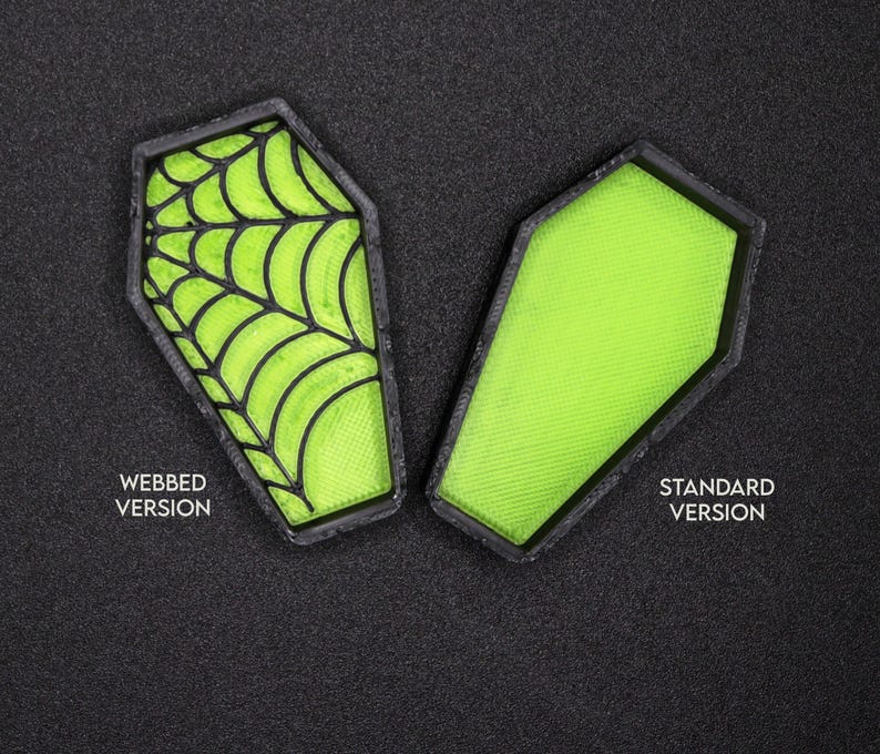 Op de afbeelding: Twee groene, kistvormige Halloween-decoraties. E&eacute;n heeft een zwart spinnenwebontwerp en de andere is effen. De tekst "WEBED VERSION" staat onder de kist met spinnenweb en "STANDARD VERSION" staat onder de effen kist.