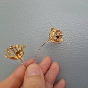 May include: A pair of gold-toned earrings shaped like open flower blossoms with thin wire stems and small circular petals. Each earring features a central cluster of stamens with tiny spherical tips and a clear silicone backing for secure attachment.