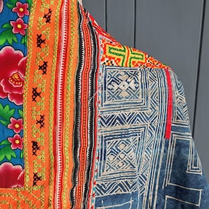 Puede incluir: Una prenda textil con un diseño de patchwork vibrante. Una sección muestra una tela azul brillante con patrones florales rojos y rosas, bordeada por paneles naranjas con bordados verdes. Otra sección muestra patrones geométricos de batik en azul y blanco.