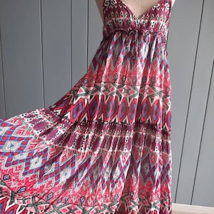 May include: A long, flowing maxi dress with a colorful geometric pattern. The dress has a V-neckline and thin straps. The pattern is a mix of red, blue, green, and black.