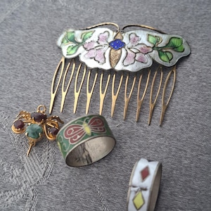 May include: A decorative hair comb with a silver-toned floral motif, a gold-toned insect-shaped brooch with red and green stones, a green band ring with a red butterfly design, and a white band ring featuring colorful diamond-shaped accents.