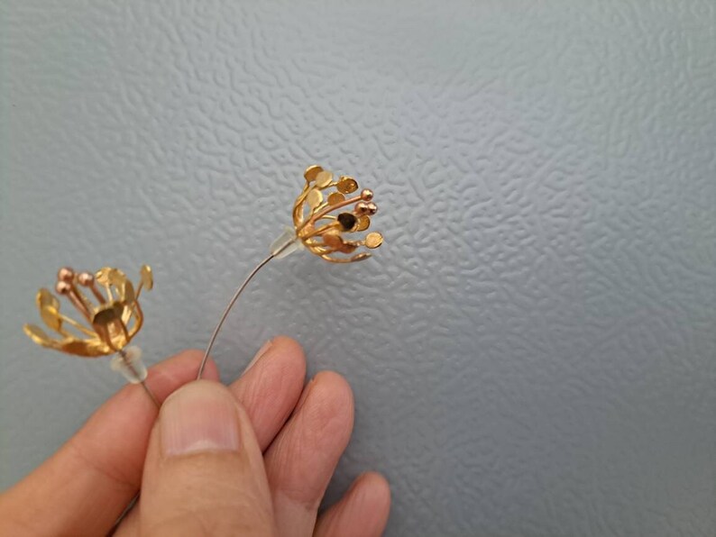 May include: A pair of gold-toned earrings shaped like open flower blossoms with thin wire stems and small circular petals. Each earring features a central cluster of stamens with tiny spherical tips and a clear silicone backing for secure attachment.