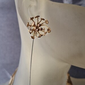 May include: A gold-toned lapel pin featuring a cluster of thin, radiating stems topped with small, flat circular discs. The design resembles a dandelion seed head or a botanical structure, with a long, straight pin extending downward from the center.