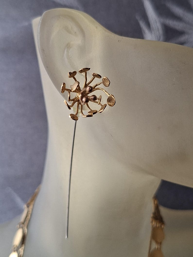 May include: A gold-toned lapel pin featuring a cluster of thin, radiating stems topped with small, flat circular discs. The design resembles a dandelion seed head or a botanical structure, with a long, straight pin extending downward from the center.