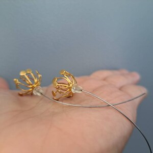 May include: A pair of gold-toned earrings featuring a spherical, cage-like structure with multiple radiating wire spokes, each tipped with a small flat disc. The earrings are attached to long, thin, curved silver-toned metal ear wires with clear backings.