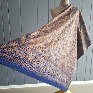 Puede incluir: Una bufanda rectangular con un color base azul profundo y un intrincado patrón floral y geométrico de color bronce dorado que se repite. La tela muestra un diseño denso y ornamentado en la superficie, terminado con un borde azul sólido.