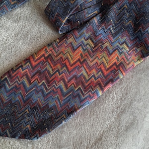 Puede incluir: Una corbata estampada con un diseño de zigzag repetido en tonos de azul, rojo, naranja y amarillo. La corbata está hecha de una tela texturizada y está sobre una superficie gris. Un accesorio clásico.