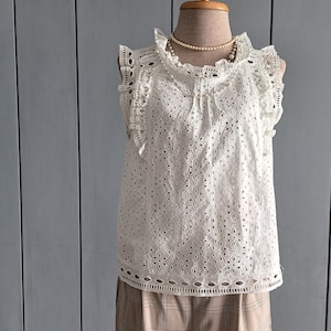 May include: White sleeveless top with eyelet detailing and a ruffled neckline. The top has a delicate, feminine design.
