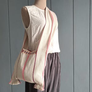 May include: A cream-colored shoulder bag made of woven fabric featuring vertical pink and dark red stripes. The bag has a wide strap, a horizontal band of geometric embroidery near the top opening, and decorative fringe along the bottom corners.