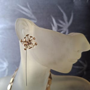 May include: A gold-toned earring featuring a cluster of small, circular discs attached to thin, radiating wires. The earring has a long, straight pin extending downward from the central cluster, creating a dandelion-like, botanical appearance.