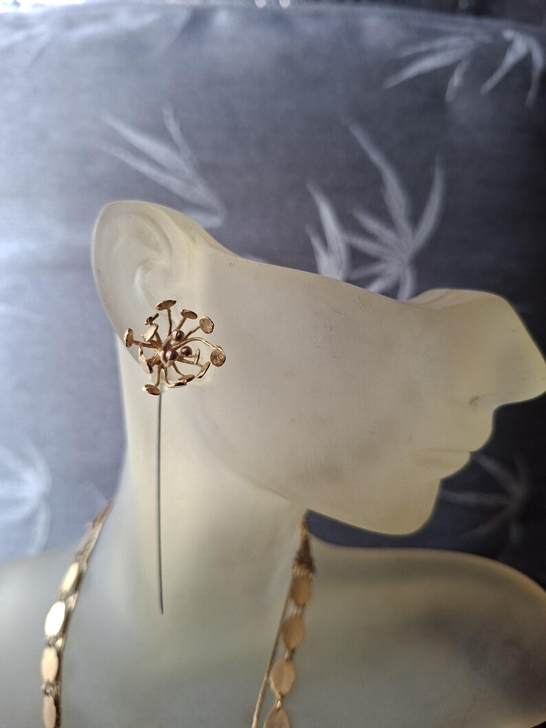 May include: A gold-toned earring featuring a cluster of small, circular discs attached to thin, radiating wires. The earring has a long, straight pin extending downward from the central cluster, creating a dandelion-like, botanical appearance.