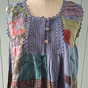 Puede incluir: Una blusa sin mangas con diseño de patchwork que presenta varios patrones florales y geométricos en tonos azul, marrón, verde y rojo. La prenda tiene un panel central de tejido de ganchillo azul con tres botones marrones y un borde tejido.