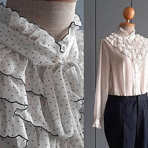 May include: A white long-sleeve blouse featuring a black polka dot pattern, a high ruffled collar, and layered scalloped ruffles across the chest with black trim. The blouse includes a front button closure and is paired with dark trousers.