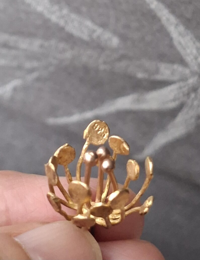 May include: A gold-colored metal jewelry piece shaped like a flower with thin stems topped by small circular discs and three central rounded stamens. One of the outer discs features the stamped text 925, indicating the metal purity of the item.