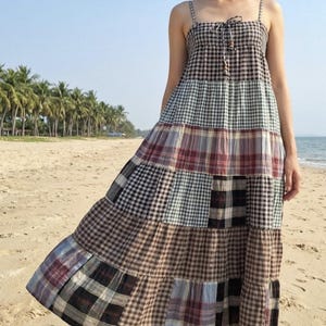 Puede incluir: Un vestido largo de niveles con un diseño de patchwork de varios patrones de cuadros y vichy en tonos marrón, negro, blanco, rojo y azul. El vestido tiene tirantes finos, un corpiño fruncido con un detalle de lazo y una falda suelta.