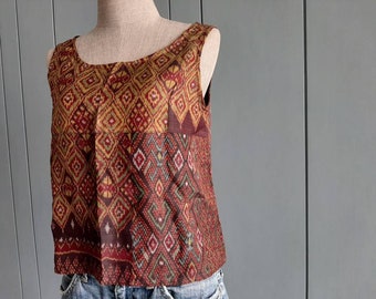 May include: A sleeveless top featuring a multi-panel design with a repeating diamond pattern in shades of deep red, mustard yellow, and dark brown. The garment has a wide scoop neckline and is worn over blue denim jeans on a mannequin torso.